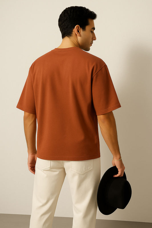 Man wearing a brown t-shirt and white pants holding a black hat against a plain background